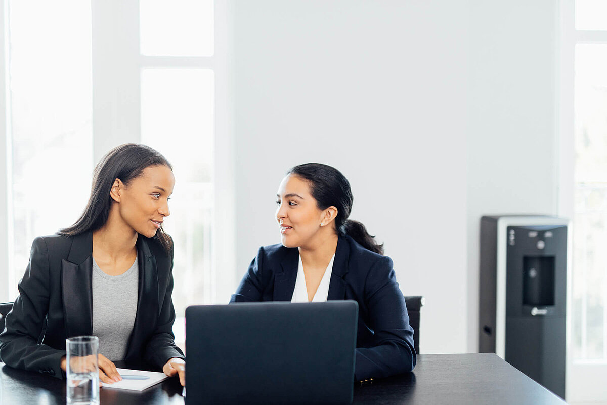 Zwei Business Frauen unterhalten sich am Arbeitsplatz mit einem Waterlogic Wasserspender im Hintergrund 